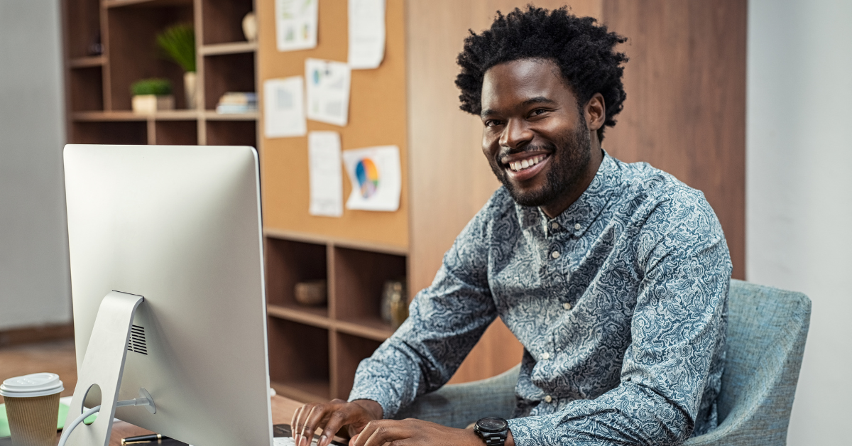 Man sitting at a desk, in front of a computer, while smiling to the camera