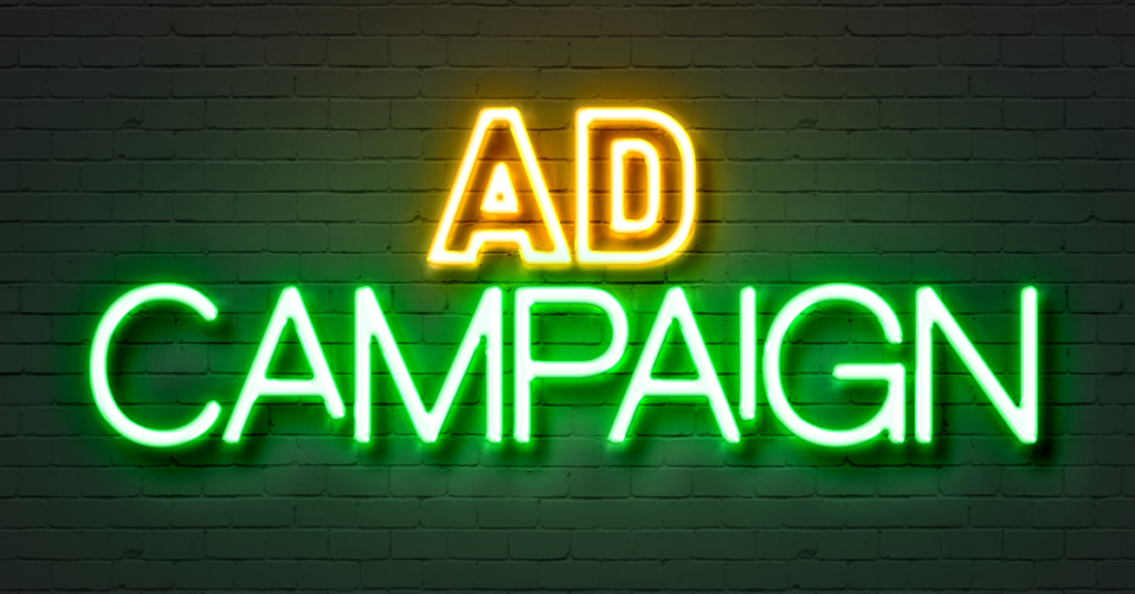 Neon sign that reads "AD CAMPAIGN" in yellow and green letters on a brick wall background.