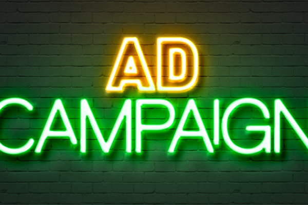 Neon sign that reads "AD CAMPAIGN" in yellow and green letters on a brick wall background.