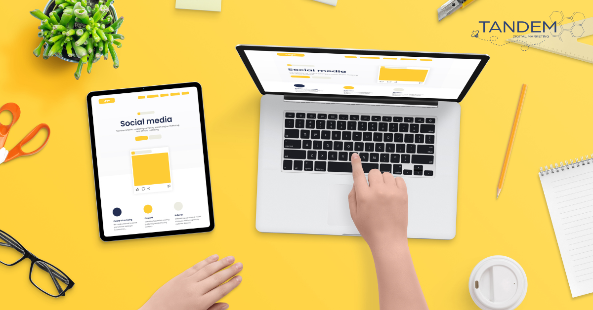 A person is working on social media content on a laptop and tablet, surrounded by office supplies on a yellow desk.