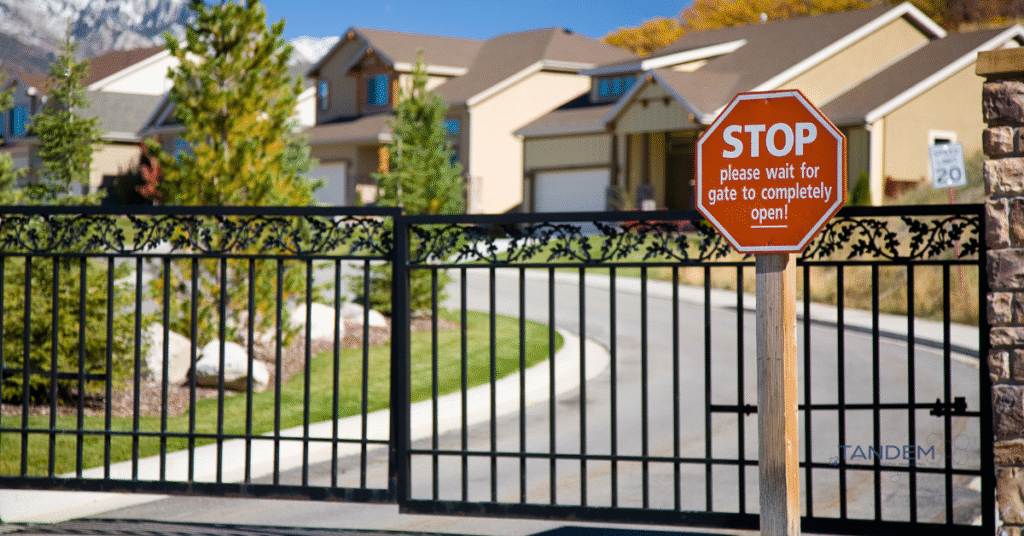 Entrance gate of a gated community with a red stop sign instructing vehicles to wait for the gate to open, with homes and landscaping in the background; Tandem Digital Marketing logo visible.