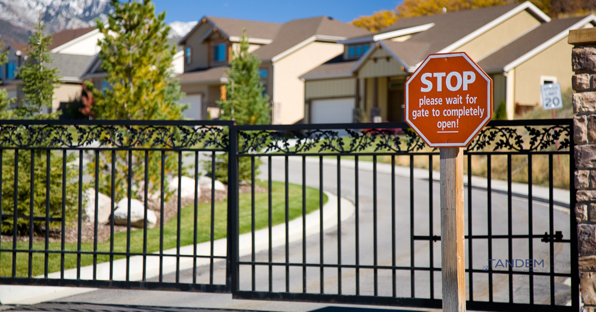 Entrance gate of a gated community with a red stop sign instructing vehicles to wait for the gate to open, with homes and landscaping in the background; Tandem Digital Marketing logo visible.