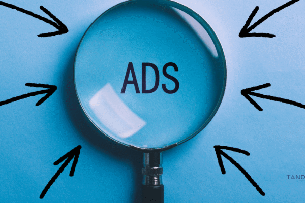 Magnifying glass highlighting the word "ADS" on a blue background with black arrows pointing inward, symbolizing ad targeting and visibility; Tandem Digital Marketing logo in the corner.