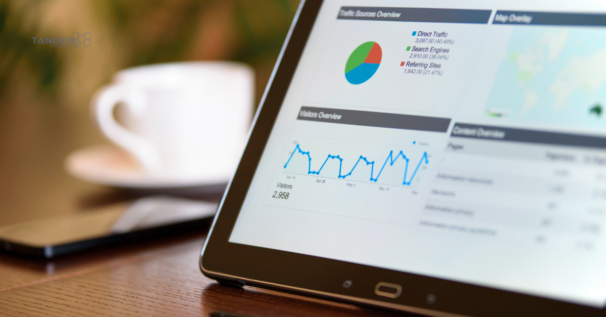 A close up of a tablet screen displaying digital marketing traffic source overview pie charts, visitor trends, and data metrics on a wooden conference table with a coffee cup in the background.