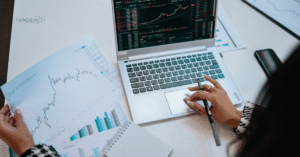 A high-angle view of a professional digital marketing strategist analyzing printed financial charts and laptop data metrics for Tandem Interactive’s August 2026 digital marketing updates.