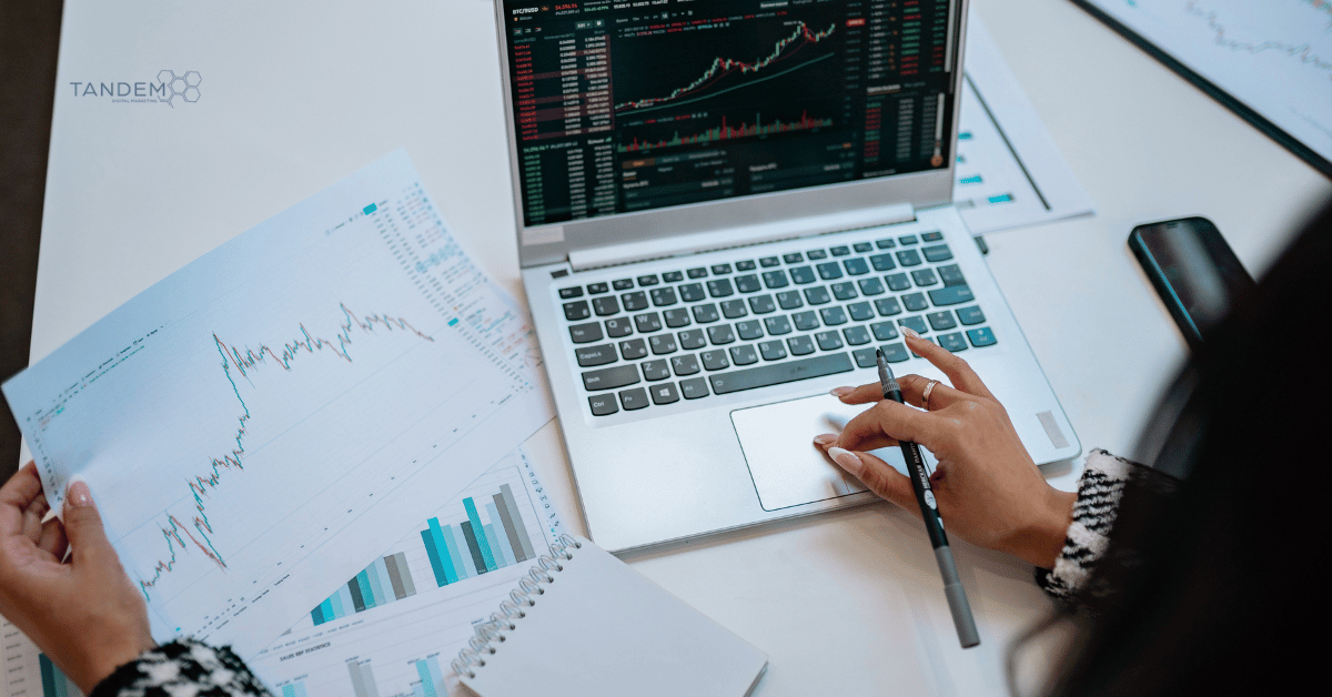 A high-angle view of a professional digital marketing strategist analyzing printed financial charts and laptop data metrics for Tandem Interactive’s August 2026 digital marketing updates.