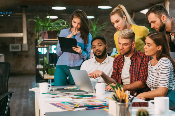A collaborative team of digital marketing strategists gathered around a laptop reviewing campaign performance metrics, featuring the new Tandem Interactive logo for our September digital marketing updates.