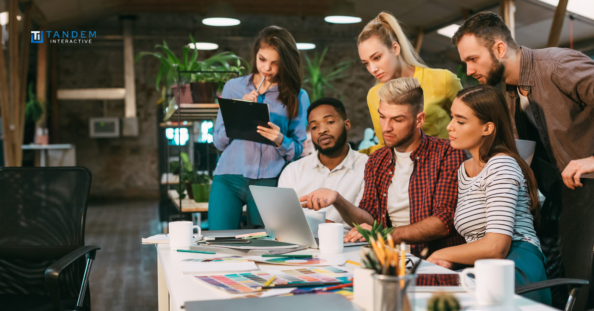 A collaborative team of digital marketing strategists gathered around a laptop reviewing campaign performance metrics, featuring the new Tandem Interactive logo for our September digital marketing updates.